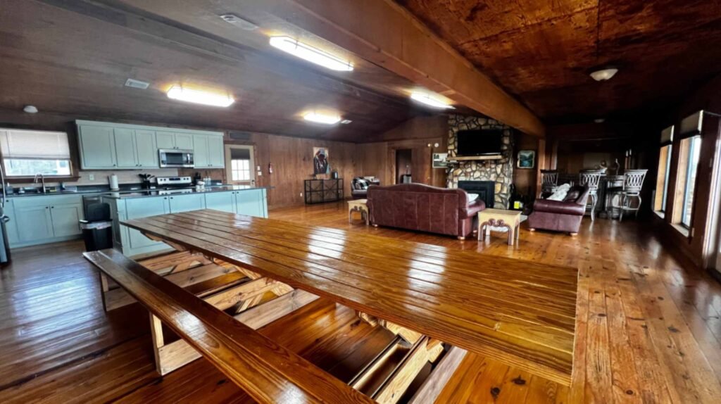 The expansive, open-concept interior of a rustic lodge at God's Country RV Park. It features warm wood-paneled walls and floors, a large picnic-style dining table, a full kitchen with light blue cabinetry, and a cozy living area with leather sofas gathered around a stone fireplace and television.
