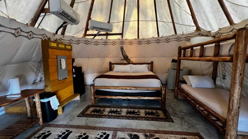 The spacious, modern interior of a glamping teepee at God's Country RV Park. It features a large queen bed with a rustic log frame, a matching wooden bunk bed, bear-themed area rugs, and wall-mounted air conditioning units for guest comfort.