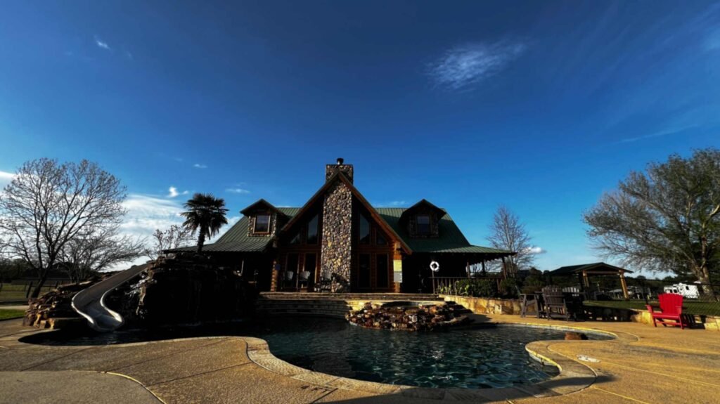 A large, rustic log lodge with a prominent stone chimney sits behind a freeform swimming pool at God's Country RV Park. The pool features a rock waterfall with a built-in slide, and the surrounding patio is dotted with a red Adirondack chair and a palm tree under a wide, blue Louisiana sky.