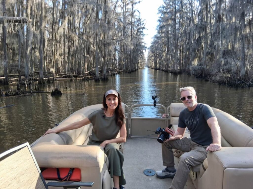 A man and woman sit on a tan pontoon boat as it travels down a narrow, straight water lane on Caddo Lake. The "boat road" is perfectly framed by towering bald cypress trees heavily draped in Spanish moss, creating a tunnel-like perspective that stretches into the distance.