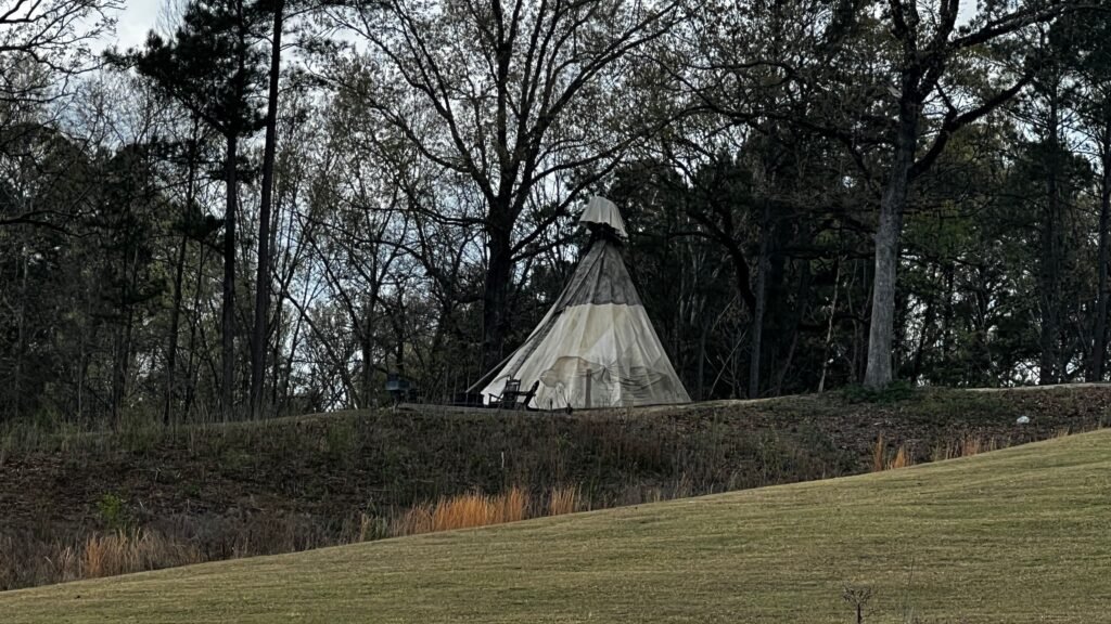 A traditional-style teepee tent sits on a grassy ridge at God's Country RV Park in Louisiana. The teepee is surrounded by a dense forest of tall, leafless trees under a clear sky, overlooking a sloping green hill.