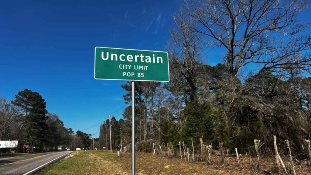 A green roadside sign against a bright blue sky reads "Uncertain City Limit, Pop 85." The sign stands next to a quiet two-lane road lined with tall pine and hardwood trees, marking the boundary of the unique Texas town on the shores of Caddo Lake.