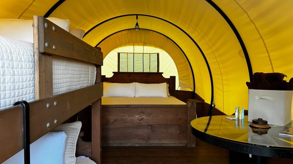 The interior of a luxury Conestoga covered wagon at God's Country RV Park. It features a bright yellow canvas arched roof, a king-sized dark wood bed frame, rustic wooden bunk beds, and a round black dining table under a single hanging Edison bulb.