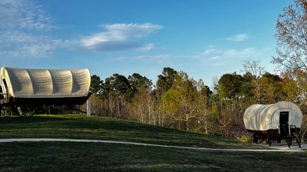 Two large, traditional-style covered Conestoga wagons sit on a grassy hillside at God's Country RV Park in Louisiana. A paved walking path curves through the green lawn, and a dense forest of pine and deciduous trees stands in the background under a clear blue sky with light clouds.