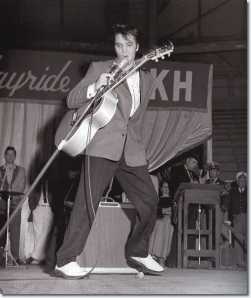 A classic black-and-white photograph of a young Elvis Presley performing on stage at the Louisiana Hayride. He is captured mid-performance, leaning back with a dynamic, energetic pose while playing an acoustic guitar and singing into a vintage silver microphone. He wears a dark blazer, light-colored slacks, and his signature white buck shoes. In the background, a large banner with "KWKH" and part of "Hayride" is visible, along with other musicians and a man at a podium.