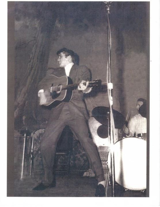 A black-and-white action photograph of a young Elvis Presley performing on stage. He is captured in a wide, energetic stance, leaning to his right while strumming an acoustic guitar. His face is turned to the side in mid-song, showing his intense focus. Behind him, a drummer is partially visible, including a bass drum and a cymbal. A tall, thin microphone stand is positioned in the foreground, framing the dynamic motion of the performance.