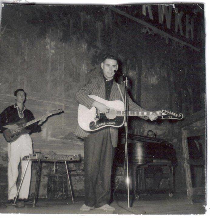 A black-and-white archival photograph of a young George Jones performing on the stage of the Louisiana Hayride. He is standing at a silver microphone, playing a large acoustic guitar with "GEORGE JONES" visible on the pickguard. He wears a striped blazer and dark trousers. Behind him to the left, a fellow musician plays an electric guitar, and a pedal steel guitar sits on a stand. In the background, a dark upright piano is visible beneath the faint "KWKH" radio station signage.