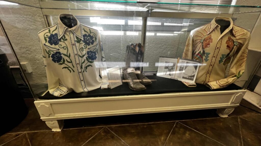 A glass museum display case inside the Shreveport Municipal Auditorium featuring iconic Western wear. On the left, a white long-sleeved shirt is adorned with vibrant blue floral embroidery. On the right, a cream-colored shirt features intricate Indigenous American profile embroidery on the chest. Between the shirts sits a pair of well-worn grey cowboy boots and an open biographical book about Hank Williams. The display is set in a white wooden base against a neutral wall.