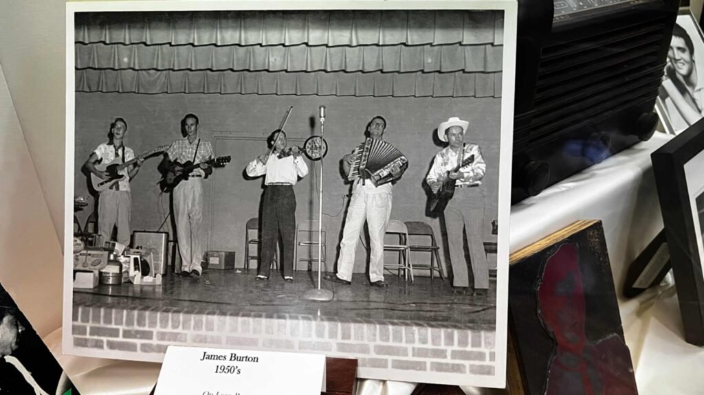 A black-and-white archival photograph displayed as part of an exhibit. The photo shows a five-piece country band performing on the brick-edged stage of the Shreveport Municipal Auditorium. On the far left, a very young James Burton plays a Fender Telecaster electric guitar. The other band members include a man on acoustic guitar, a fiddle player, an accordionist, and a man in a cowboy hat playing a hollow-body guitar. A small white label below the photo reads "James Burton."