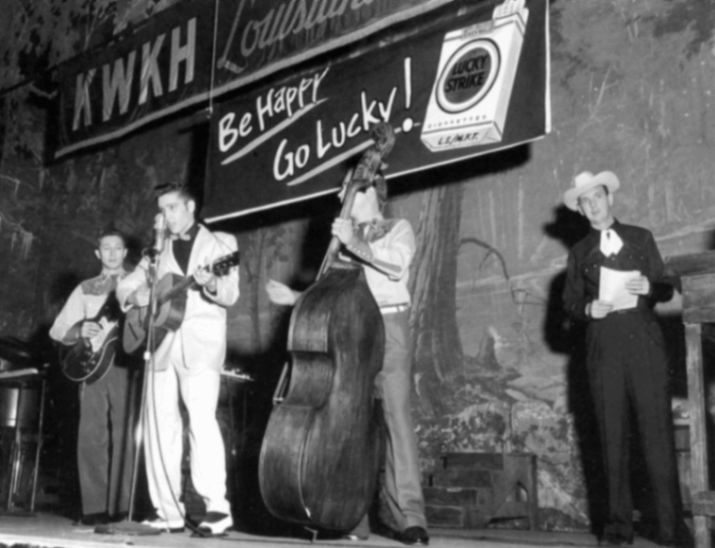 A historic black-and-white photograph of Elvis Presley performing on the Louisiana Hayride stage at the Shreveport Municipal Auditorium. Elvis, in a light-colored suit, sings into a vintage microphone and plays guitar. He is accompanied by Scotty Moore on electric guitar and Bill Black on upright bass. To the right, an announcer in a cowboy hat and western wear stands at a podium holding a script. A large "Lucky Strike" cigarette advertisement banner with the slogan "Be Happy Go Lucky!" hangs prominently above the performers next to the KWKH station sign.