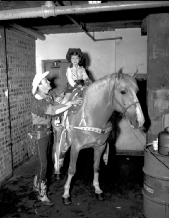 A black-and-white historical photograph taken in a backstage or stable area of the Shreveport Municipal Auditorium. "King of the Cowboys" Roy Rogers, dressed in full fringed western regalia and a white cowboy hat, smiles as he holds a young girl securely on the back of his famous palomino horse, Trigger. The young girl wears a cowboy hat and a western shirt, smiling brightly at the camera. The background shows industrial elements like brick walls, exposed pipes, and a metal barrel, highlighting the behind-the-scenes setting.
