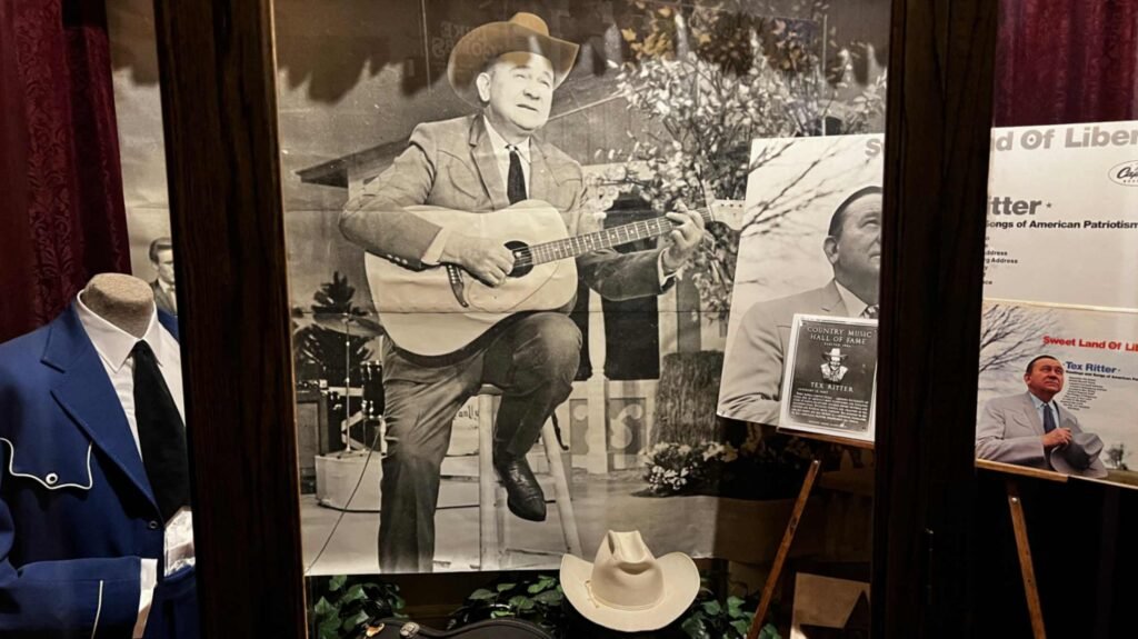 An indoor exhibit at the Shreveport Municipal Auditorium dedicated to Tex Ritter. The display features a large black-and-white mural of Ritter in a cowboy hat playing an acoustic guitar. To the left, a headless mannequin wears a sharp blue Western suit with a black tie. A white cowboy hat sits at the base of the mural. On the right, a smaller display features his Country Music Hall of Fame plaque and an album cover for "Sweet Land of Liberty," reinforcing his legacy as a patriotic American icon.
