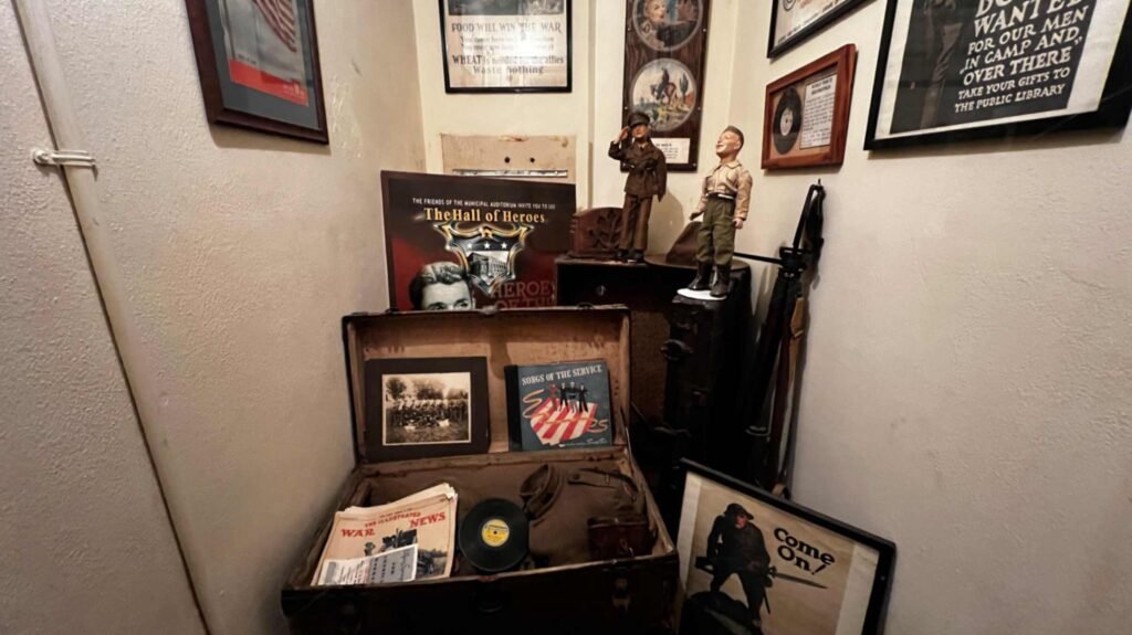 A corner museum display titled "The Hall of Heroes" inside the Shreveport Municipal Auditorium, featuring World War I and II era military memorabilia. A large open vintage trunk is filled with items like a "War News" newspaper, a "Songs of the Service" record, and a gas mask. Above the trunk, two military figurines stand on a dark chest. The walls are covered in framed period posters, including one that reads "Food Will Win the War" and another recruiting for the public library. A poster for "The Hall of Heroes" featuring a soldier's face is also prominent.