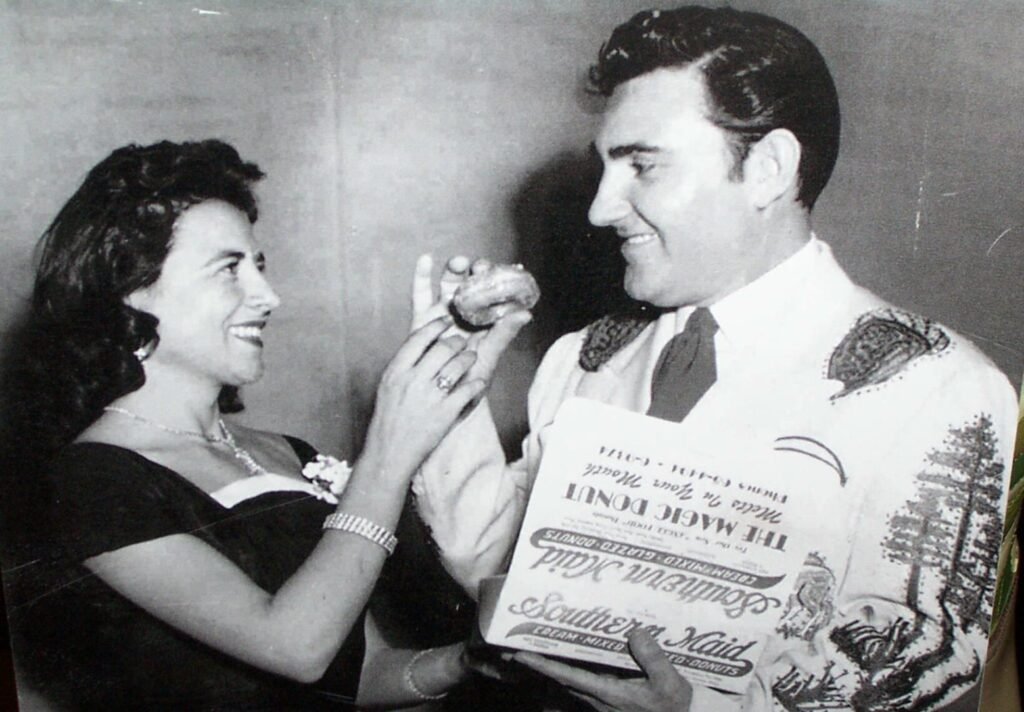 A black-and-white promotional photograph featuring a young, smiling Elvis Presley and a dark-haired woman. Elvis is wearing a light-colored Western shirt with embroidered details. The woman is holding a glazed donut up to Elvis’s mouth while he holds an open box of "Southern Maid Donuts," which features the slogan "The Magic Donut - Melts in Your Mouth."