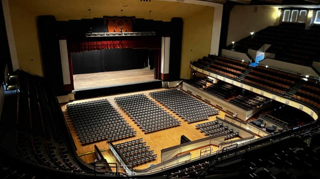 A high-angle, wide-perspective interior shot of the Shreveport Municipal Auditorium from the upper balcony. The vast wooden main floor is neatly arranged with hundreds of black folding chairs facing a large, historic stage. The stage is framed by an ornate Art Deco proscenium arch with red velvet curtains and white fluted columns. The theater features a deep, horseshoe-shaped balcony with multi-level seating that wraps around the entire room, all under a high, dimly lit ceiling.