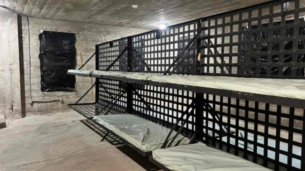An interior view of a communal cell block in the DeRidder Gothic Jail featuring rows of metal-framed bunk beds with thin mattresses attached to a wall of thick iron latticework.