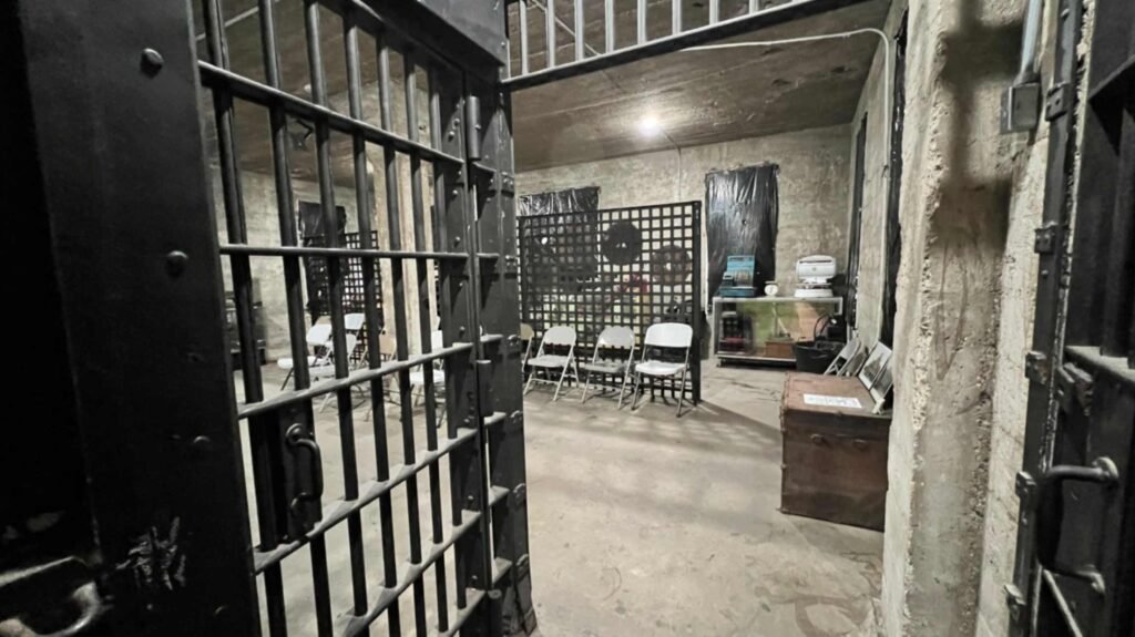 A view through heavy, open iron bars into a dim cell block room where white folding chairs are set up for a tour among historical trunks and artifacts.