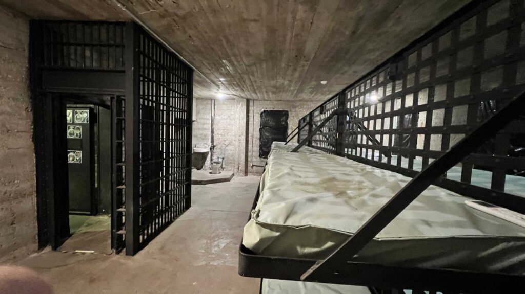 An interior view of a dim cell block at the DeRidder Gothic Jail featuring rows of metal bunk beds with thin mattresses and a black iron-barred partition.