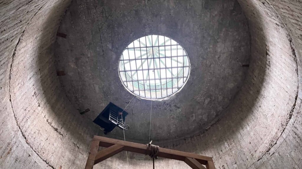 A low-angle view looking straight up a circular concrete shaft toward a barred skylight, with the wooden beam of a gallows and a hanging rope visible in the foreground.