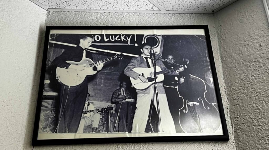 A black-and-white photograph in a black frame, hanging on a textured white wall. The photo shows a young Elvis Presley performing on the Louisiana Hayride stage. He is front and center, singing into a vintage microphone and playing an acoustic guitar. To his left, Scotty Moore plays a hollow-body electric guitar, and to his right, Bill Black is visible with his upright bass. In the background, a portion of a "Lucky Strike" banner is visible at the top of the frame.
