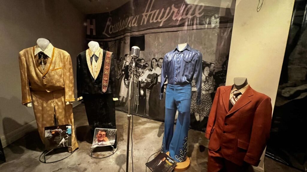 An exhibit inside the Shreveport Municipal Auditorium featuring four headless mannequins displaying diverse stage outfits against a large black-and-white mural of a "Louisiana Hayride" performance. From left to right: a shimmering gold patterned suit with a matching bow tie, a black tuxedo-style jacket with a gold vest and a decorated guitar strap, a bright blue satin long-sleeved shirt with matching bell-bottom trousers featuring decorative silver embroidery at the cuffs, and a classic burnt-orange two-piece suit with a striped tie. A vintage silver microphone on a stand is positioned in the center foreground.