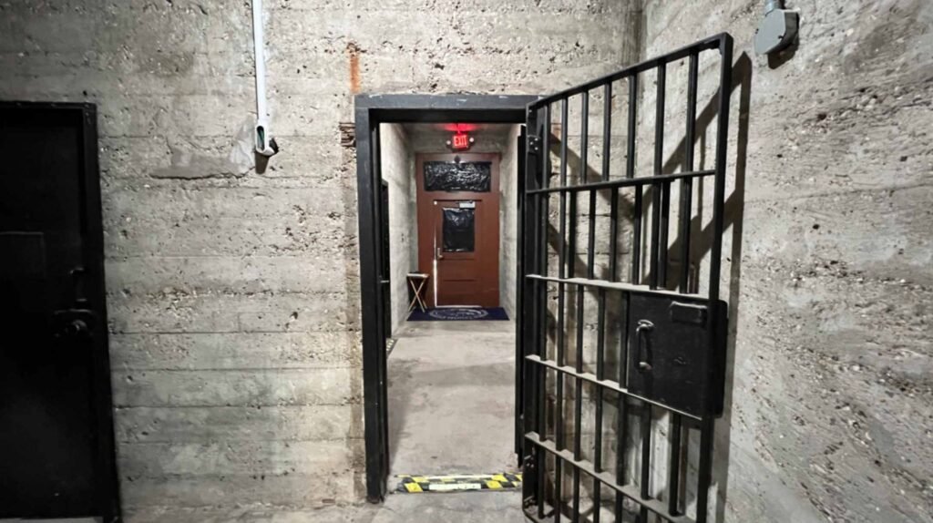A heavy iron jail cell door stands open, revealing a hallway with a modern wooden door and a red "EXIT" sign at the end of a concrete corridor in the DeRidder Gothic Jail.