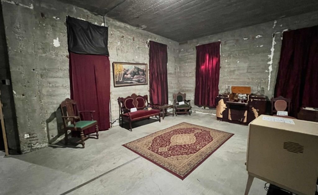 An interior room inside the DeRidder Gothic Jail, staged with an ornate red rug and vintage Victorian-style furniture against raw, unfinished concrete walls and dark curtains.