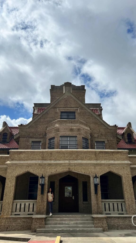 A low-angle, full-front view of the historic Beauregard Parish Jail in DeRidder, Louisiana. The tan, stone-textured Gothic Revival building features multiple gables with red-tiled roofs, barred windows, and a central arched entrance where a person is standing. A dramatic, cloudy blue sky sits behind the towering structure.