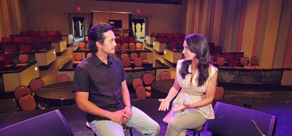 Musician Laine Hardy and host Karen LeBlanc are seated on barstools on the edge of the Teche Theater stage, facing each other in conversation. Laine is on the left, looking intently at Karen as she speaks with expressive hand gestures. Behind them, the empty theater seats and tables are visible, bathed in a soft, atmospheric purple glow.
