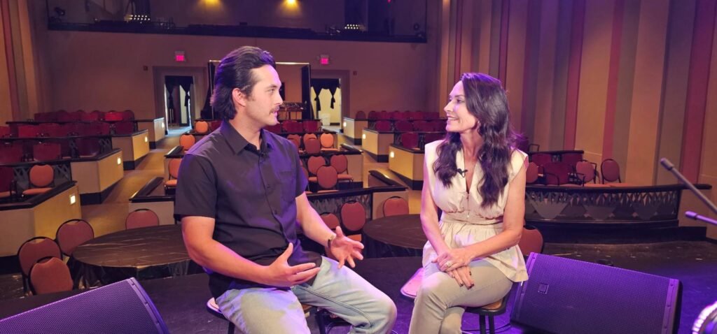 Laine Hardy is seated on a stool on the Teche Theater stage, speaking earnestly to host Karen LeBlanc. He gestures with his hands as he shares a story, while Karen looks on with an appreciative smile. The historic theater’s empty seating and warm purple lighting create an intimate atmosphere for their conversation.