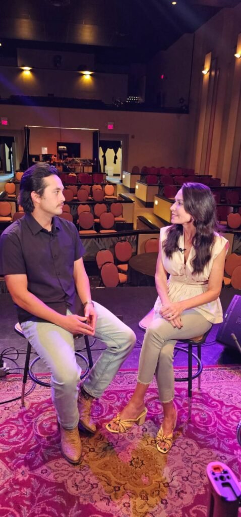 A full-length shot of Laine Hardy and host Karen LeBlanc seated on tall stools for their LA64 interview. They are positioned on an ornate, red-patterned rug on the Teche Theater stage. Laine wears a black short-sleeved button-down, light-wash jeans, and tan cowboy boots, while Karen is in a cream peplum top, grey trousers, and gold sandals. The empty theater seats extend into the background under warm, ambient lighting.