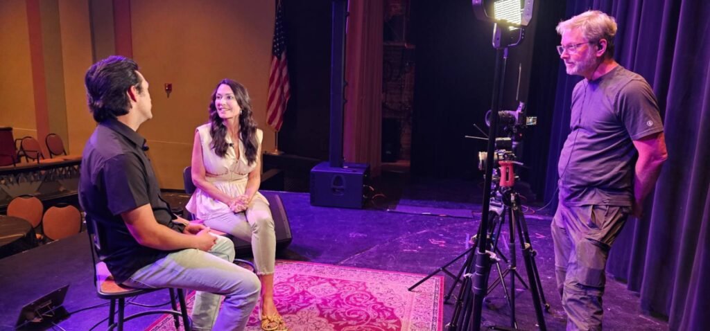An wide shot on the stage of the historic Teche Theater showing host Karen LeBlanc and musician Laine Hardy seated for an interview. They are positioned on a traditional rug under theatrical lighting. To the right, a production crew member stands behind a professional camera on a tripod, capturing the conversation. The background features dark stage curtains and an American flag.