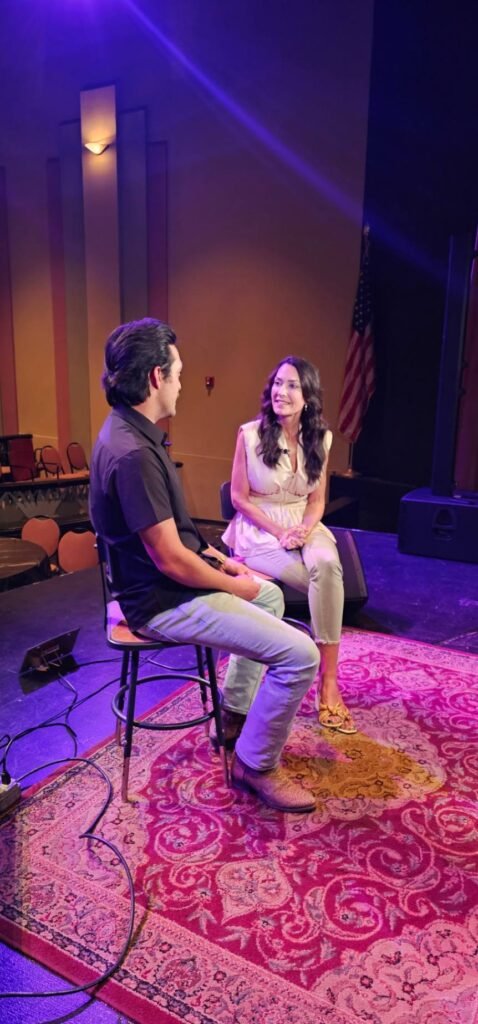 Host Karen LeBlanc and Laine Hardy are captured in a candid moment during their interview on the Teche Theater stage. Seated on tall stools atop an ornate Persian-style rug, they are engaged in conversation under vibrant purple and warm yellow stage lights. Laine is seen from the side, wearing a black shirt and light jeans, while Karen looks toward him, smiling.
