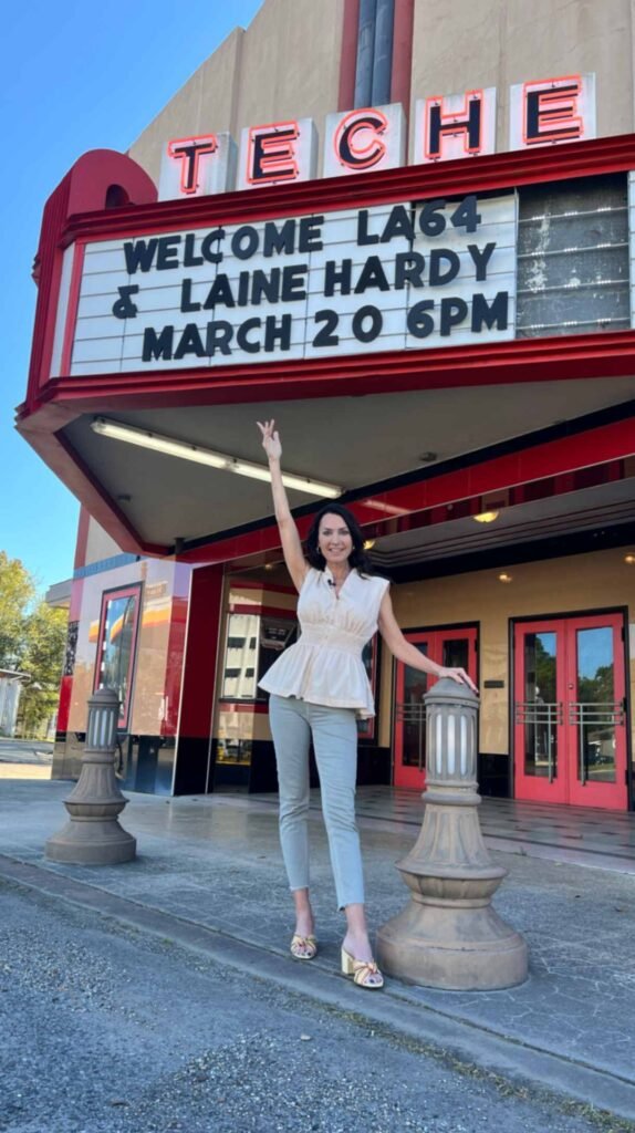 Karen stands in front of the historic Teche Theater in Franklin, Louisiana. She is wearing a light-colored peplum top and grey trousers, with one arm raised toward the theater’s vintage marquee. The marquee reads, "WELCOME LA64 & LAINE HARDY MARCH 20 6PM" in bold black lettering.