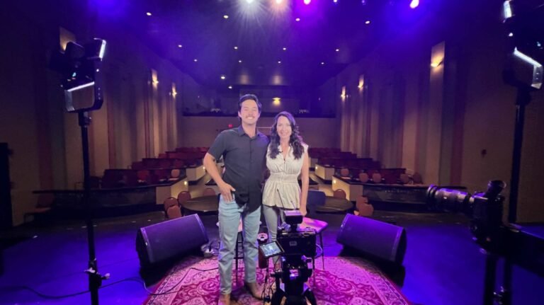 Laine Hardy and the interviewer stand together on the stage of the dimly lit Teche Theater. They are positioned on an ornate rug, surrounded by stage monitors and filming equipment under purple lighting. The empty theater seats are visible in the background, creating a quiet, expectant atmosphere.