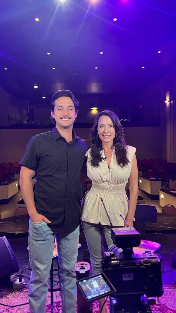 A portrait of host Karen LeBlanc and musician Laine Hardy standing together on the stage of the Teche Theater. Karen is wearing a light-colored peplum top, and Laine is in a black short-sleeved button-down. The camera equipment in the foreground and the purple-hued theater seating in the background capture the behind-the-scenes essence of their LA64 interview.