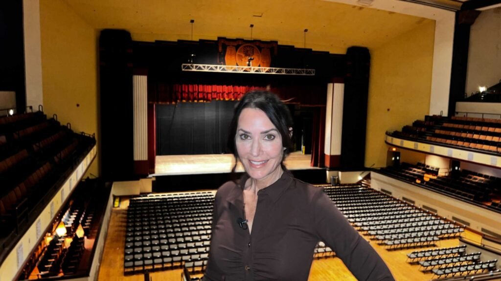 A woman with dark hair and a brown shirt smiles in the foreground of a high-angle shot overlooking the interior of the Shreveport Municipal Auditorium. Behind her, the massive theater opens up, showing the main floor filled with rows of black chairs and the historic stage in the distance. The stage is framed by an ornate Art Deco proscenium arch with red curtains and white fluted columns, highlighting the immense size of the venue.