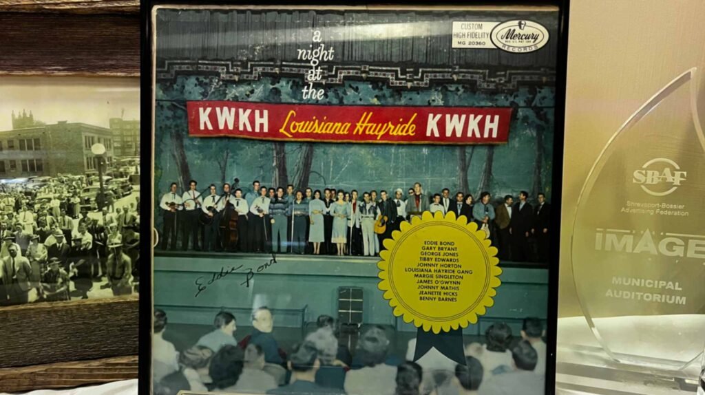 A close-up of a framed "A Night at the KWKH Louisiana Hayride" vinyl record album cover displayed inside the Shreveport Municipal Auditorium. The cover art features a colorful photograph of a large group of musicians and singers standing on the auditorium's stage before a live audience. A bright yellow starburst sticker on the cover lists featured performers, including George Jones and Johnny Horton. The album is flanked by a historic black-and-white photo of a crowd and a modern glass "IMAGE" award for the Municipal Auditorium.