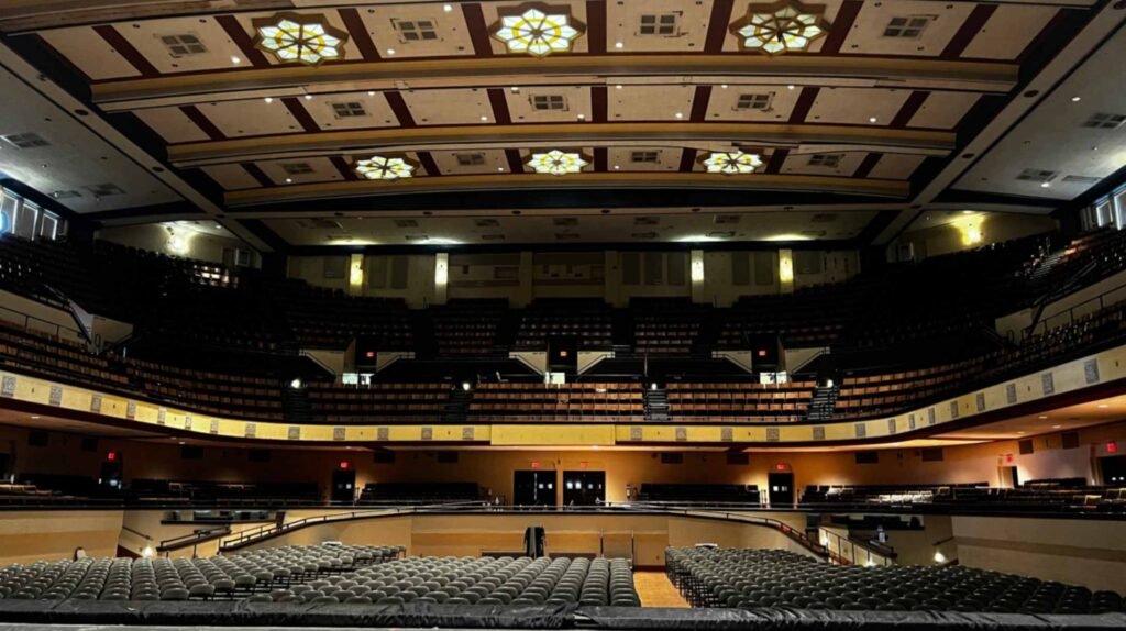 A wide-angle interior view from the stage of the Shreveport Municipal Auditorium, looking out toward the vast seating area. The image shows the main floor filled with rows of chairs, a large curved balcony, and upper gallery seating under a high, ornate ceiling. The ceiling features decorative rectangular panels and several large, glowing star-shaped light fixtures that illuminate the darkened hall with a warm, golden glow.
