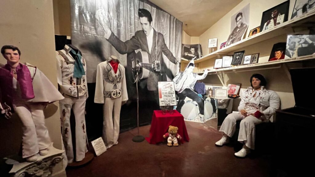 A vibrant interior room within the Shreveport Municipal Auditorium dedicated to Elvis Presley memorabilia. The room features several life-sized mannequins wearing iconic Elvis-style jumpsuits, including a white "eagle" design. A large black-and-white backdrop shows a young Elvis performing. Shelves are lined with framed photographs, awards, and various collectibles. A life-sized figure of Elvis in a white jumpsuit sits in a chair to the right, and a small teddy bear sits on a red-clothed pedestal in the center.