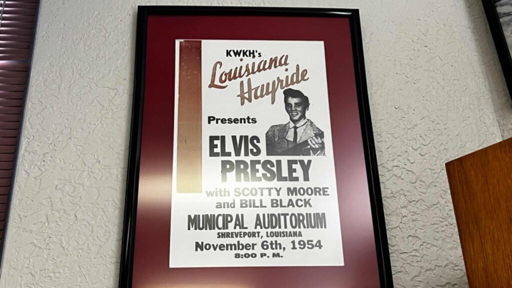 A framed vintage promotional poster for "KWKH's Louisiana Hayride" hanging on a textured wall. The poster, set against a deep maroon mat in a black frame, features a black-and-white photo of a young, smiling Elvis Presley. The text prominently advertises "ELVIS PRESLEY with SCOTTY MOORE and BILL BLACK" at the "MUNICIPAL AUDITORIUM, SHREVEPORT, LOUISIANA." The date is clearly listed as "November 6th, 1954" at "8:00 P.M."