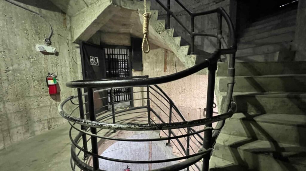 A high-angle view looking down into the center of a circular concrete spiral staircase with a replica hanging noose suspended from a beam inside the DeRidder Gothic Jail.