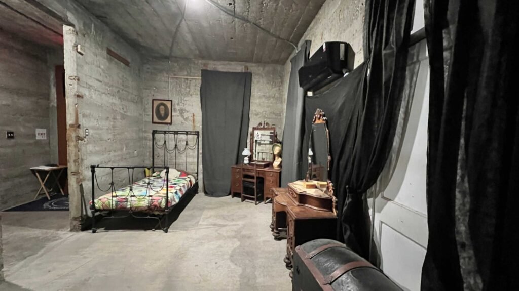 An interior room inside the DeRidder Gothic Jail, staged with a vintage metal bed frame, a wooden vanity, and black drapes against cold, unfinished concrete walls.