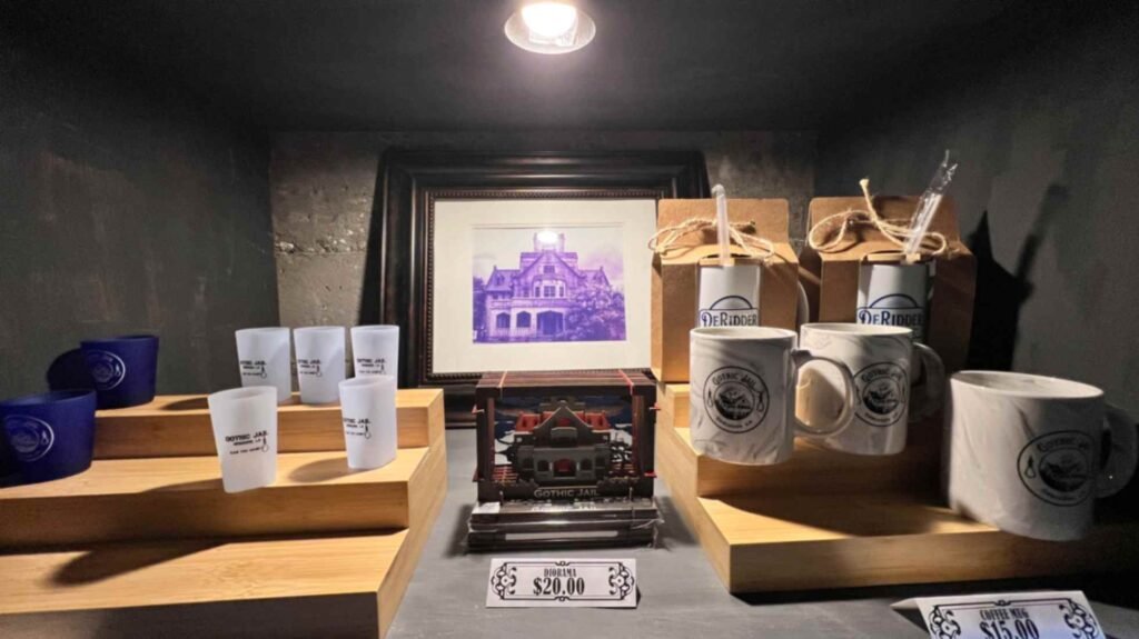 A display shelf in the DeRidder Gothic Jail gift shop featuring branded mugs, shot glasses, a diorama, and a framed photo of the jail's exterior under a single spotlight.