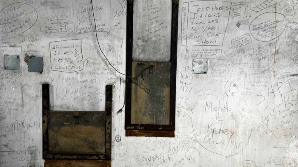 A close-up photograph of a weathered, white-painted backstage wall inside the Shreveport Municipal Auditorium, covered in decades of handwritten graffiti, names, and dates from performers and stagehands. Notable inscriptions include "HAYRIDE 1/28/67," "EARL GRANT SHOW," and "JEFF HARRIS." The wall features old metal electrical boxes, exposed wiring, and two dark rectangular openings or access panels, all surrounded by layers of scribbled history.