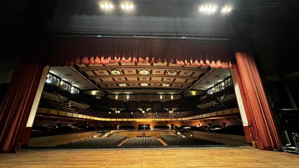 A dramatic wide-angle shot from the back of the wooden stage at the Shreveport Municipal Auditorium, looking through the proscenium arch. Heavy red velvet curtains are pulled back, framing the vast, darkened theater. The warm glow of the auditorium's star-shaped ceiling lights and theater lamps illuminates the empty rows of seating and the multi-level balconies, creating a sense of anticipation and history.