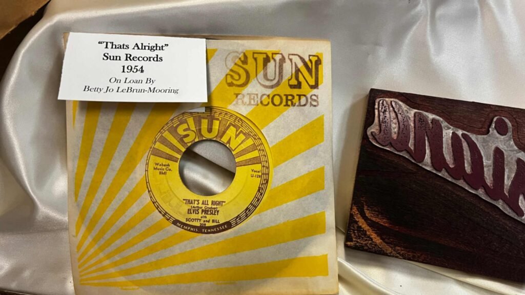 A close-up shot of a rare 1954 original Sun Records 45rpm vinyl record of Elvis Presley’s first single, "That’s All Right." The record features the iconic yellow-and-brown Sun Records label and is housed in its original yellow-and-white sunburst paper sleeve. A small white card pinned to the sleeve identifies the record and notes it is "On Loan By Betty Jo LeBrun-Mooring." To the right, a portion of a wooden block with the word "Muni" carved in stylized script is visible on a white satin background.