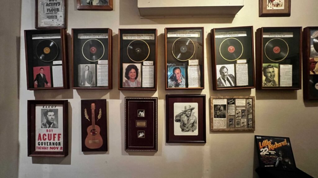 A museum-style gallery wall inside the Shreveport Municipal Auditorium featuring a collection of framed memorabilia honoring country music legends. The top row contains several gold and platinum records alongside portraits of artists like Slim Whitman and Faron Young. The bottom rows feature framed posters and photographs, including a "Roy Acuff for Governor" campaign poster, an illustration of a guitar, and a drawing of Hank Williams. A "Little Richard" album cover is visible in the lower-right corner.