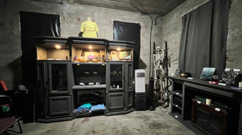 A dimly lit gift shop area inside the DeRidder Gothic Jail featuring a large gray entertainment center used as a display shelf, a skeleton prop on a ladder, and concrete walls.