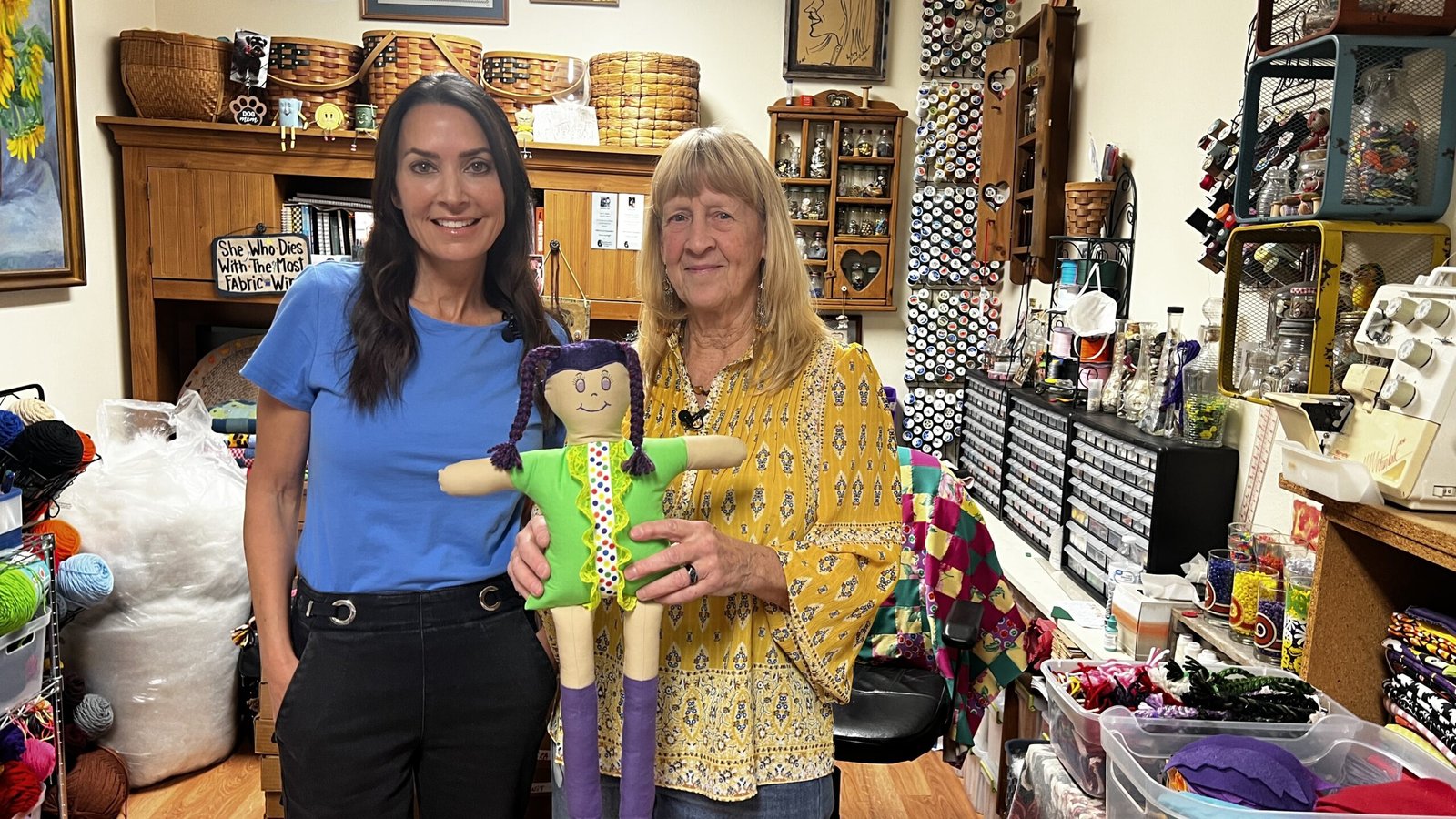 Karen and Anne holding a handmade rag doll together in a craft room filled with sewing supplies.